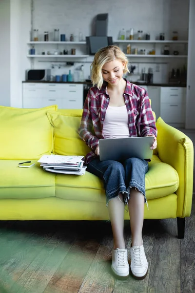 Усміхнена блондинка в картатій сорочці працює з дому з ноутбуком на колінах — стокове фото