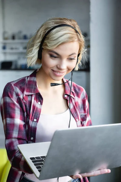 Mujer rubia joven con camisa a cuadros y auriculares trabajando con el ordenador portátil - foto de stock