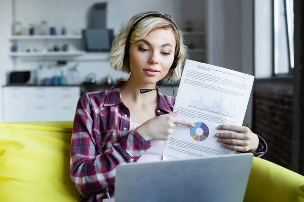 Jeune femme blonde dans les écouteurs ayant conférence en ligne et montrant des graphiques — Photo de stock