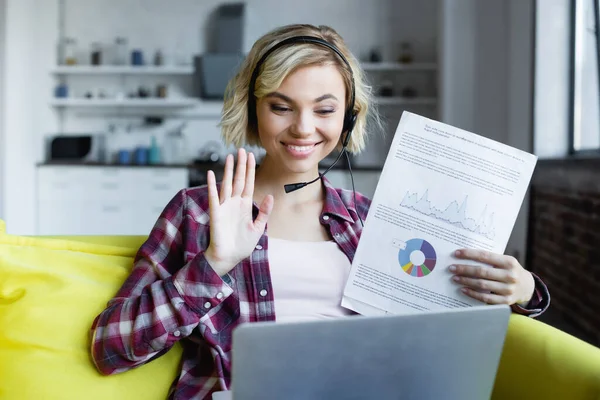Young blonde woman in headphones showing graphs and waving hand — Stock Photo