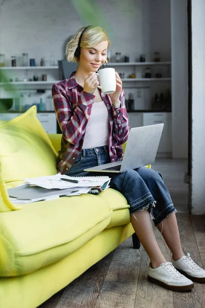 Young blonde woman in headphones working from home and drinking tea — Stock Photo