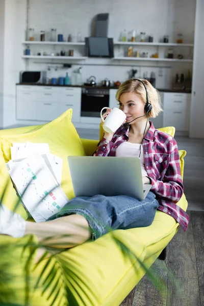 Joven rubia en auriculares trabajando desde casa y bebiendo té - foto de stock