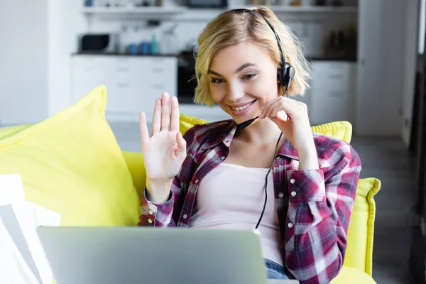 Jeune femme blonde dans les écouteurs ayant conférence en ligne et agitant la main — Photo de stock