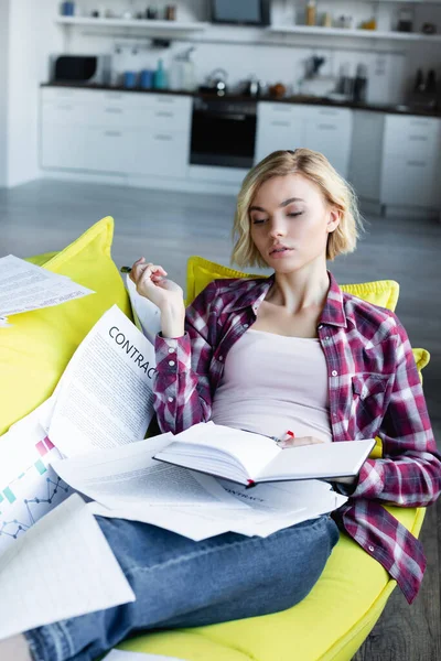 Jovem loira em camisa quadriculada deitada no sofá com documentos e segurando notebook — Fotografia de Stock