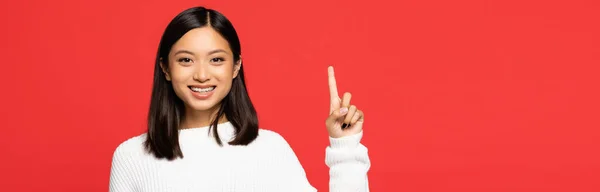 Happy asian woman in sweater pointing with finger isolated on red, banner — Stock Photo