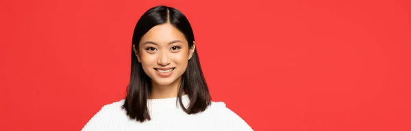 Happy and young asian woman in sweater looking at camera isolated on red, banner — Stock Photo