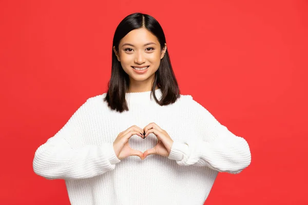 Happy and young asian woman showing heart sign with hands isolated on red — Stock Photo