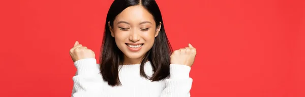 Happy and young asian woman with closed eyes rejoicing isolated on red, banner — Stock Photo