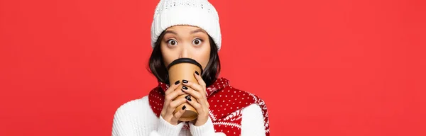 Здивована азіатська жінка в капелюсі і шарфі п'є каву, щоб піти ізольовано на червоному, банер — стокове фото