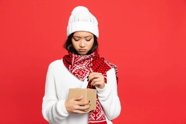 Triste asiatique femme dans chapeau et écharpe tenant boîte de tissu isolé sur rouge — Photo de stock