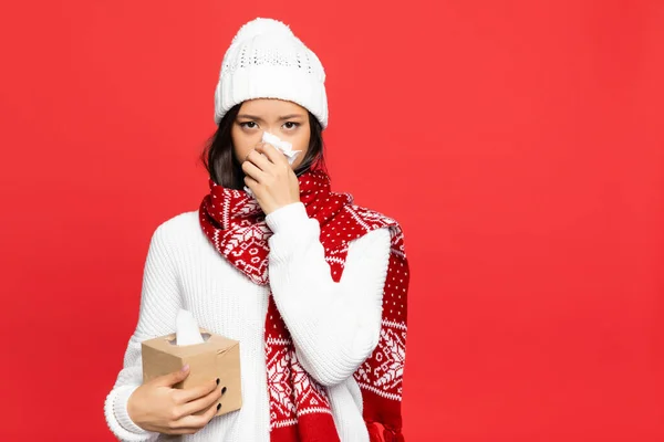 Хвора азіатка в капелюсі і шарфі тримає серветку і тканинну коробку, чхаючи ізольовано на червоному — стокове фото