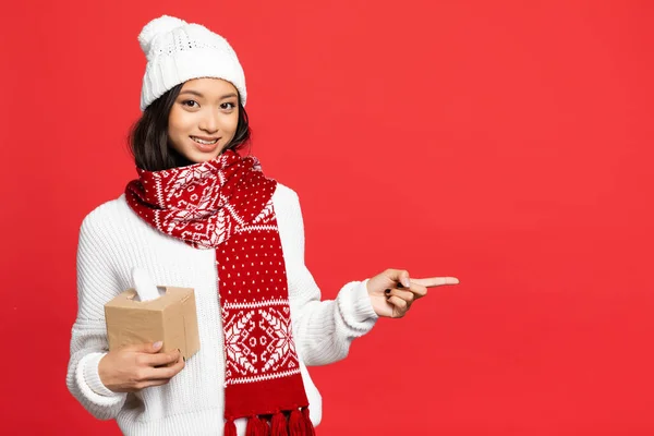 Feliz e jovem mulher asiática em chapéu e cachecol segurando caixa de tecido e apontando com o dedo isolado no vermelho — Fotografia de Stock