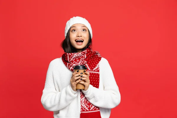 Excited asian woman in hat and scarf holding disposable cup with coffee to go isolated on red — Stock Photo