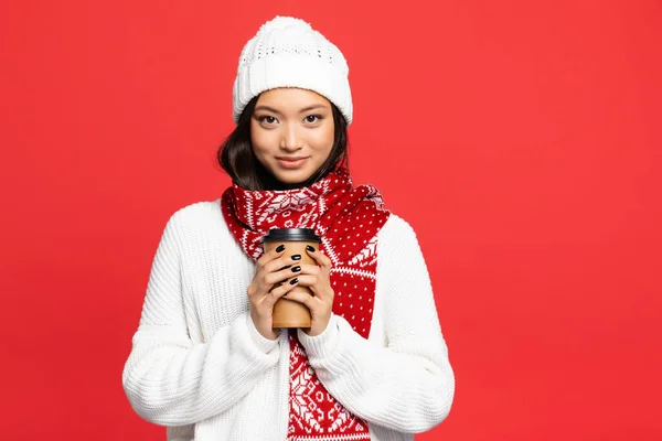 Mujer asiática feliz en sombrero y bufanda sosteniendo taza desechable con café para ir aislado en rojo - foto de stock