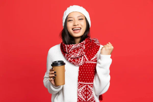 Eccitato donna asiatica in cappello e sciarpa tenendo tazza usa e getta e gioire isolato su rosso — Foto stock