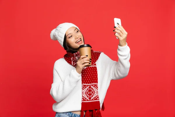 Excitada mujer asiática en sombrero y bufanda sosteniendo taza desechable y tomando selfie aislado en rojo - foto de stock