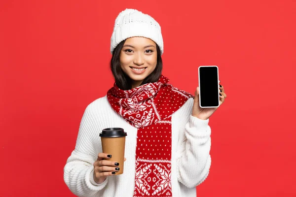 Glücklich asiatische Frau mit Hut und Schal hält Smartphone mit leerem Bildschirm und Kaffee to go auf rot — Stockfoto