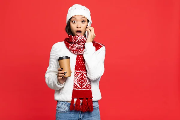 Conmocionado mujer asiática en sombrero y bufanda celebración de café para ir mientras habla en el teléfono inteligente aislado en rojo - foto de stock