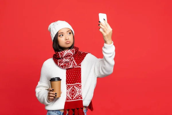 Disgustado mujer asiática en sombrero y bufanda sosteniendo café para ir mientras toma selfie aislado en rojo - foto de stock