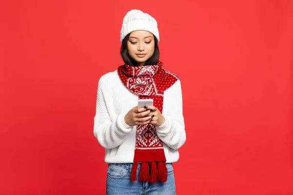 Joven asiático mujer en sombrero y bufanda mensajes de texto en smartphone aislado en rojo - foto de stock