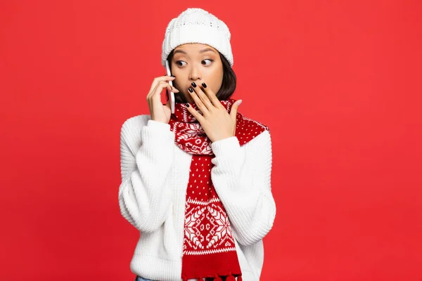 Sorprendido asiático mujer en sombrero y bufanda hablando en smartphone y cubriendo boca aislado en rojo - foto de stock