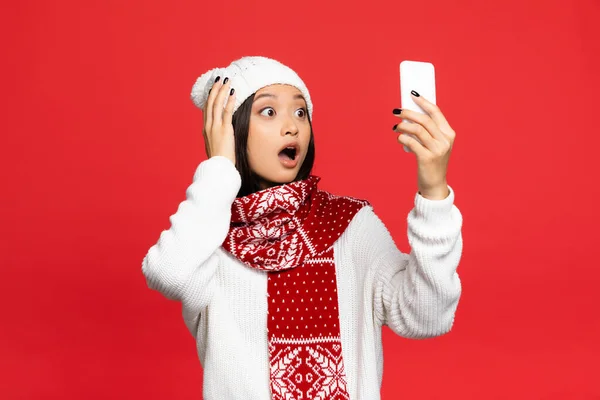 Sorpreso asiatico donna in cappello e sciarpa holding smartphone mentre prende selfie isolato su rosso — Foto stock