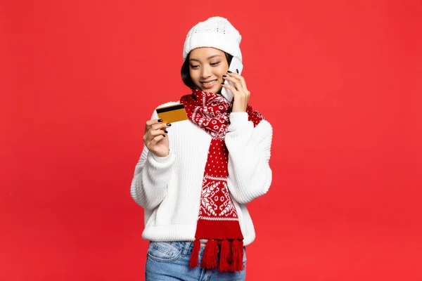 Sonriente asiático mujer en sombrero y bufanda celebración smartphone y mirando a la tarjeta de crédito aislado en rojo - foto de stock