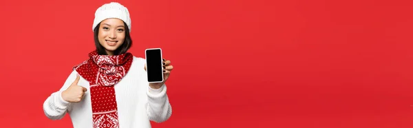 Улыбающаяся азиатская женщина в шляпе и шарфе, держащая смартфон с пустым экраном, показывая большой палец, изолированный на красном, баннер — стоковое фото