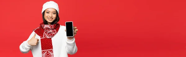 Sonriente asiático mujer en sombrero y bufanda celebración smartphone con pantalla en blanco, mientras que muestra el pulgar hacia arriba aislado en rojo, bandera - foto de stock