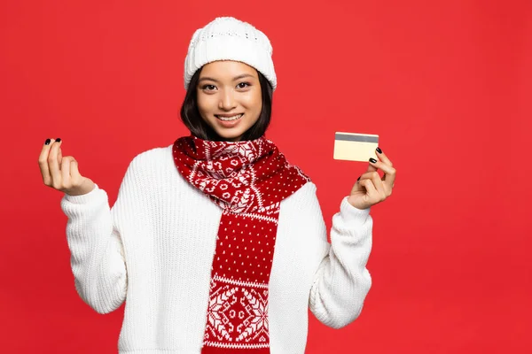 Lächelnde asiatische Frau mit Hut und Schal hält Kreditkarte in der Hand und zeigt Geldgeste auf rotem Hintergrund — Stockfoto