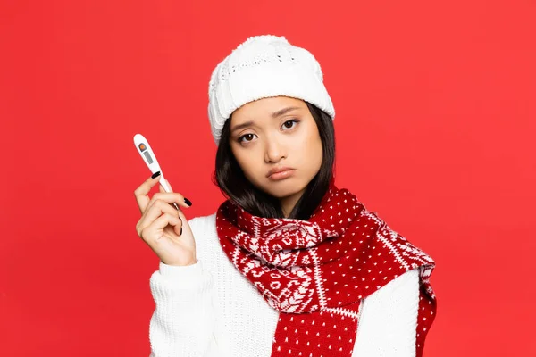 Sad asian woman in hat and scarf holding electronic thermometer isolated on red — Stock Photo