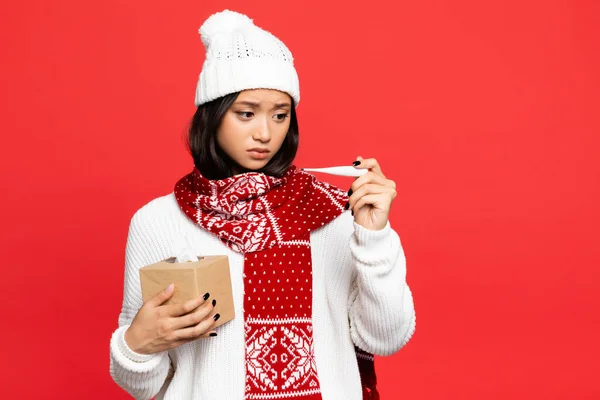 Mujer asiática enferma en sombrero y bufanda sosteniendo termómetro electrónico y caja de tejido aislado en rojo - foto de stock