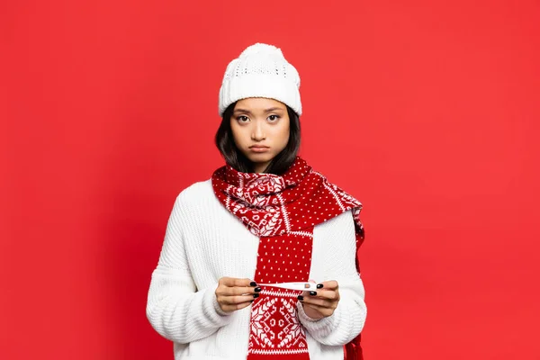 Upset and sick asian woman in hat and scarf holding electronic thermometer isolated on red — Stock Photo
