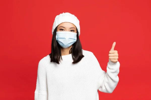 Joven asiático mujer en médico máscara y sombrero mostrando pulgar arriba aislado en rojo - foto de stock