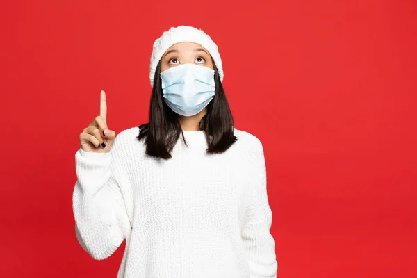 Young asian woman in medical mask and hat pointing with finger while looking up isolated on red — Stock Photo