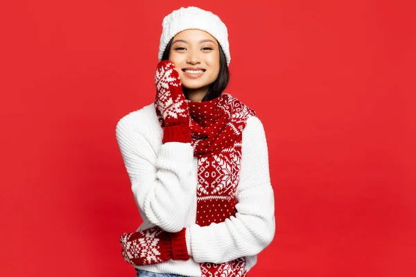 Cheerful young asian woman in white hat, mittens and scarf smiling isolated on red — Stock Photo