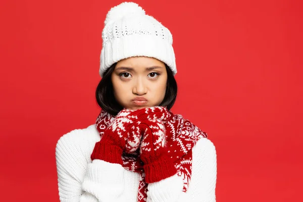 Sad asian woman in white hat, mittens and scarf looking at camera and pouting lips isolated on red, banner — Stock Photo