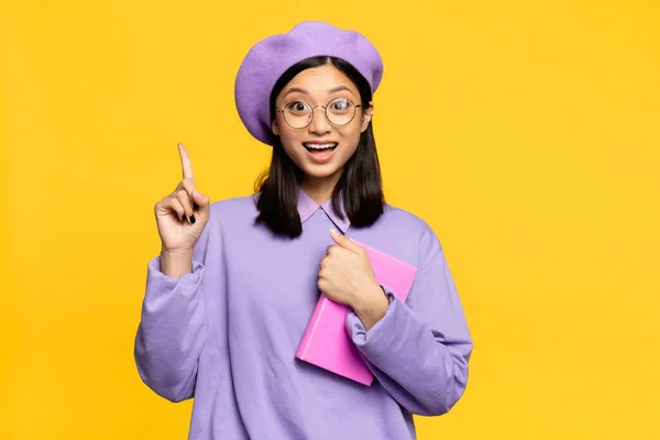 Excited asian woman in beret and eyeglasses holding notebook and pointing with finger isolated on yellow — Stock Photo