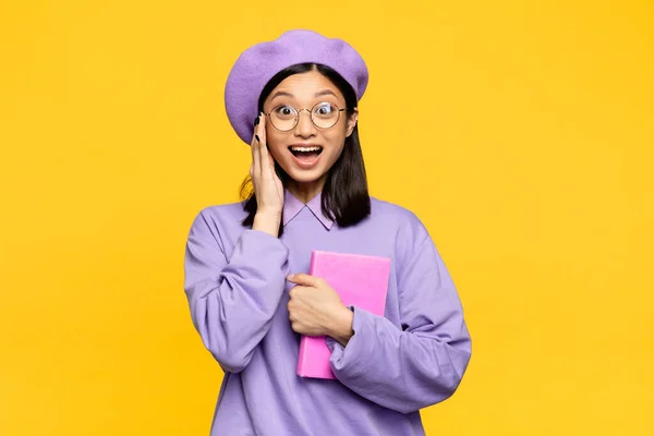 Aufgeregte asiatische Frau in Baskenmütze mit Notizbuch und Einstellbrille isoliert auf gelb — Stockfoto