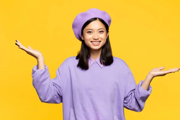 Felice donna asiatica in berretto puntando con le mani isolate sul giallo — Foto stock