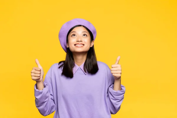 Feliz asiático mulher no boina mostrando polegares para cima e olhando para cima isolado no amarelo — Fotografia de Stock