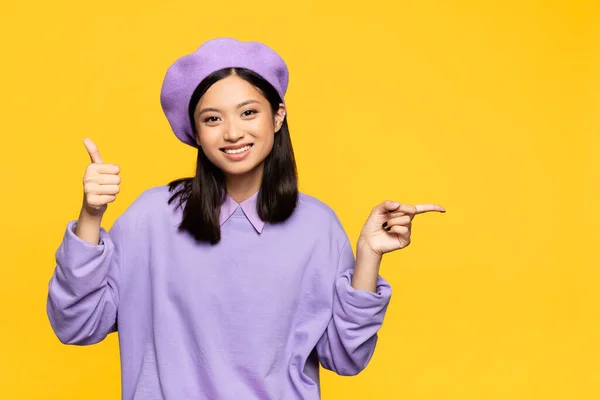 Happy asian woman in beret showing thumb up and pointing with finger isolated on yellow — Stock Photo