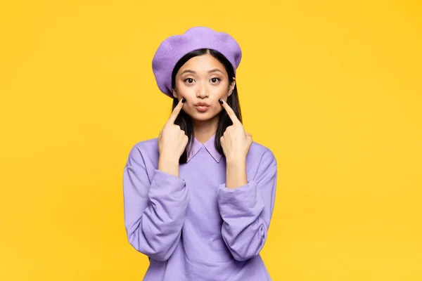 Asian woman in beret touching cheeks with fingers while pouting lips isolated on yellow — Stock Photo