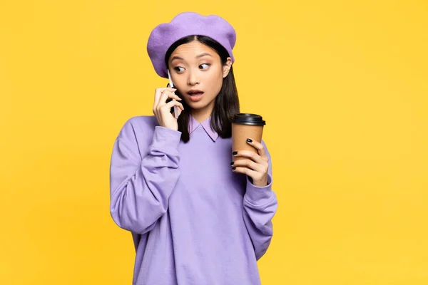 Surpresa asiático mulher no boina segurando café para ir e falando no smartphone isolado no amarelo — Fotografia de Stock