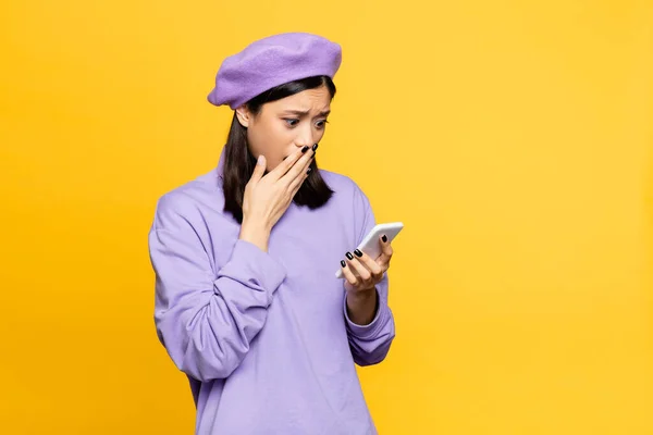 Triste asiatique femme dans béret regarder smartphone et couvrant bouche isolé sur jaune — Photo de stock