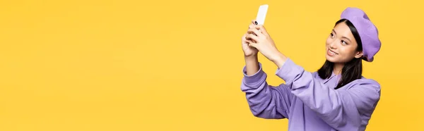 Feliz asiático mujer en boina tomando selfie en smartphone aislado en amarillo, bandera - foto de stock