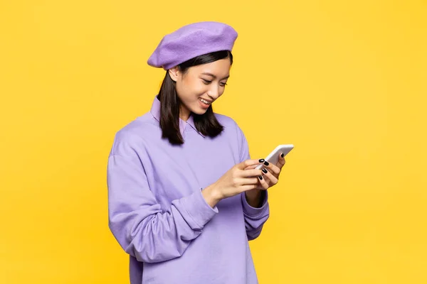 Feliz asiático mulher no boina olhando para smartphone isolado no amarelo — Fotografia de Stock