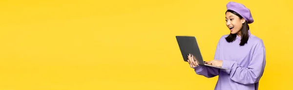 Happy asian woman in beret using laptop isolated on yellow, banner — Stock Photo