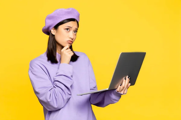 Pensive asian woman in beret using laptop isolated on yellow — Stock Photo