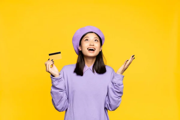 Excitada mujer asiática en boina celebración de tarjeta de crédito y mirando hacia arriba aislado en amarillo - foto de stock